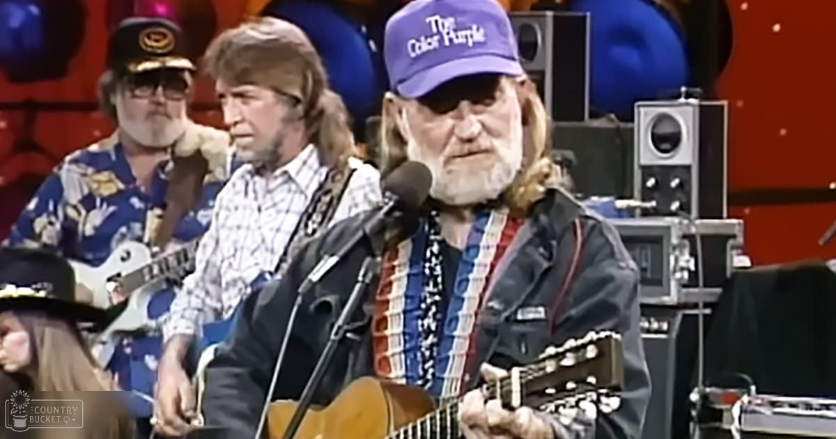 Adult male musician wearing a blue baseball cap with 'The Color Purple' printed on it, black leather jacket over patterned shirt, and American flag lanyard, playing an acoustic guitar in a music studio or stage setting.