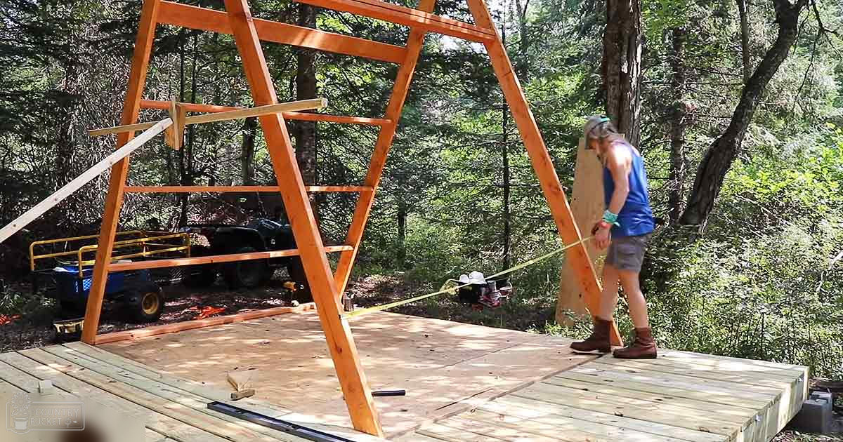 Adult female constructing wooden frame in wooded outdoor area with planks, tape measure, and utility trailer visible.