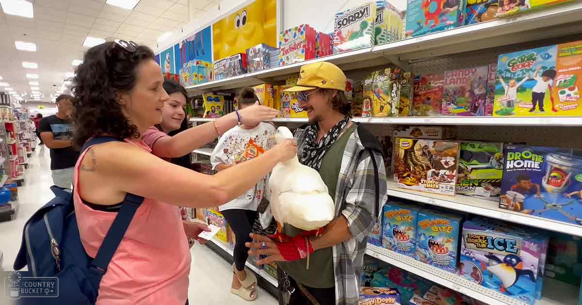 An adult female in a pink top holds a toddler with colorful designs in her arms while interacting with another adult male holding a white stuffed animal inside a brightly lit toy section of a store, surrounded by shelves stocked with board games and toys.