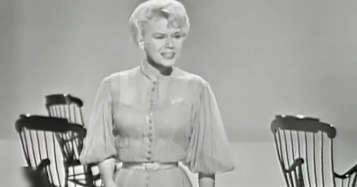 Adult female in a mid-20th-century blouse and skirt set with loose, bell-shaped sleeves, standing indoors against a plain light-colored wall.