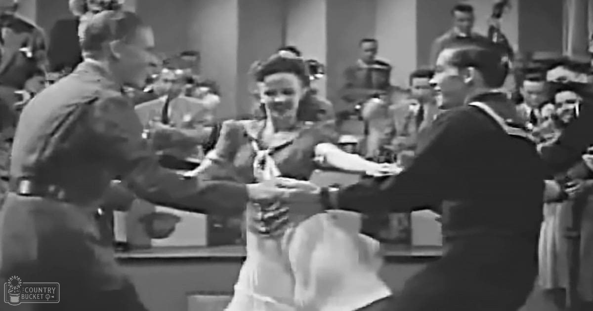 A social dance scene with two young adults dancing closely together in a formal setting from an earlier era, surrounded by other individuals in attire suggesting a social gathering.
