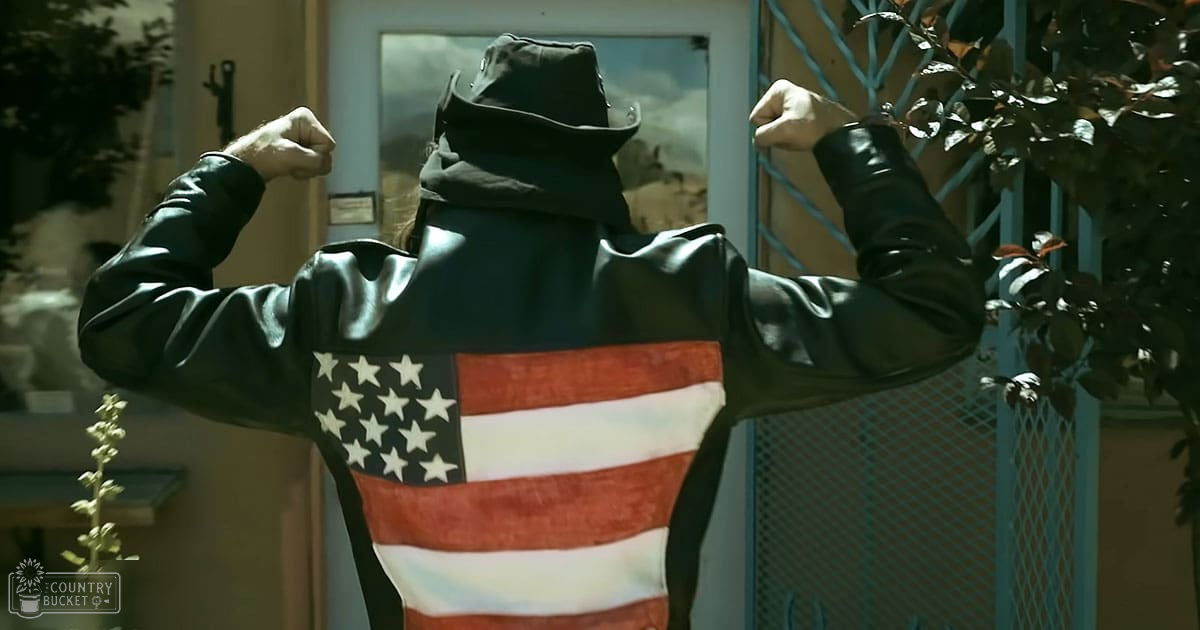 An adult male wearing a black, American flag-themed hooded jacket made of leather or synthetic material stands in front of a house with a door and some plants. The scene is set during daytime, possibly late afternoon.