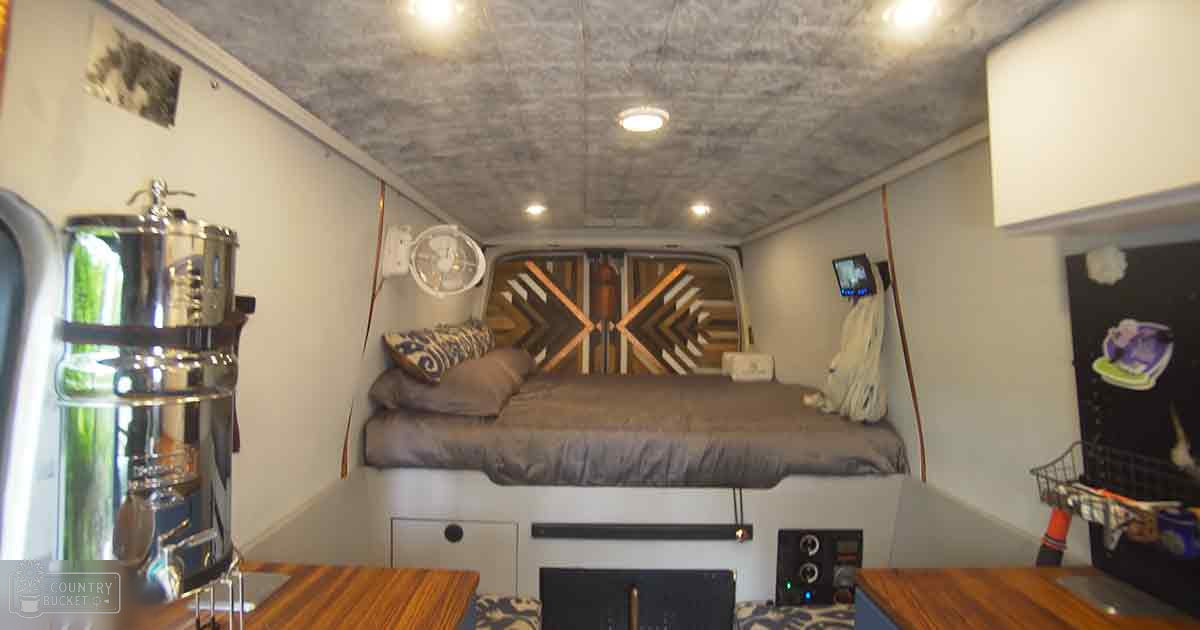 Converted van interior, patterned bedding, metallic container, wooden table, television screen, black refrigerator with magnets, small stove or heater.