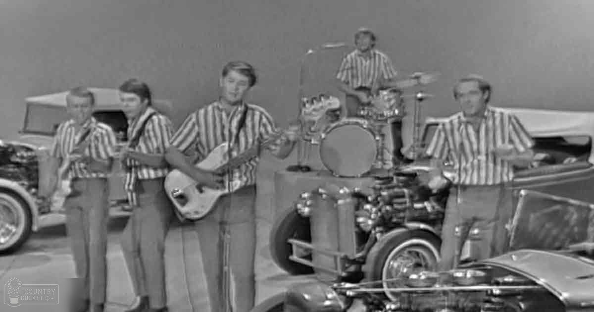 A musical group performing with vintage cars as props against a plain studio backdrop.