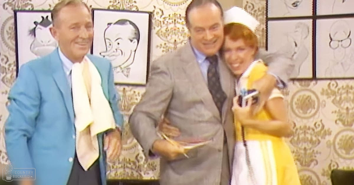 A group photo taken indoors with decorative wallpaper, with three adults: a male in a light blue blazer over white shirt and dark trousers; another male in a gray suit jacket, patterned tie, and dark trousers holding a book or folder; and a female in a yellow dress with white stripes on sleeves and skirt, wearing a white hat with a ribbon. The setting is indoors with even daytime lighting.