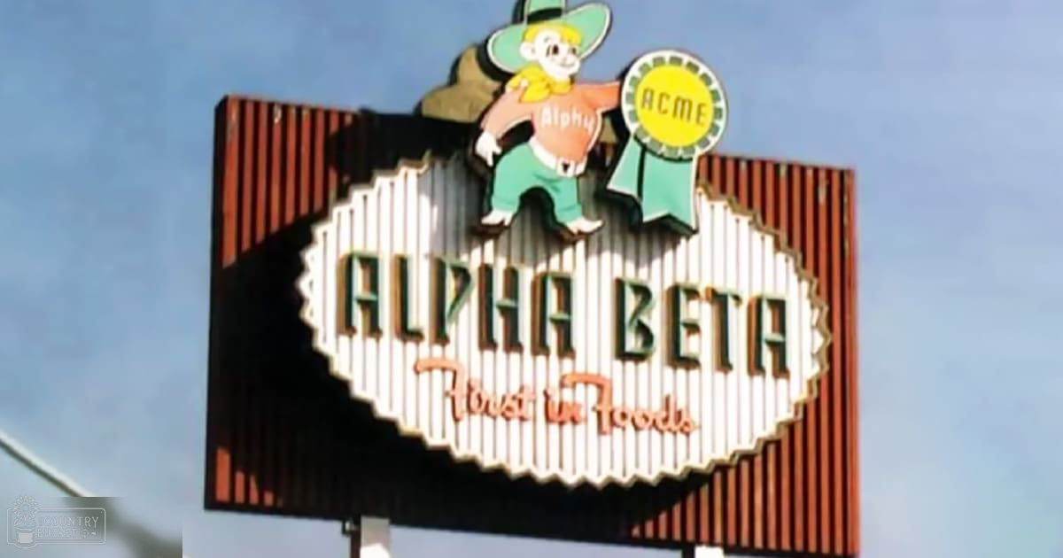An outdoor scene with a sign for 'Alpha Beta Foods' with a cartoon character named 'Alphy' holding an ACME sack. The setting is bright, clear sky under daylight in summer or spring.
