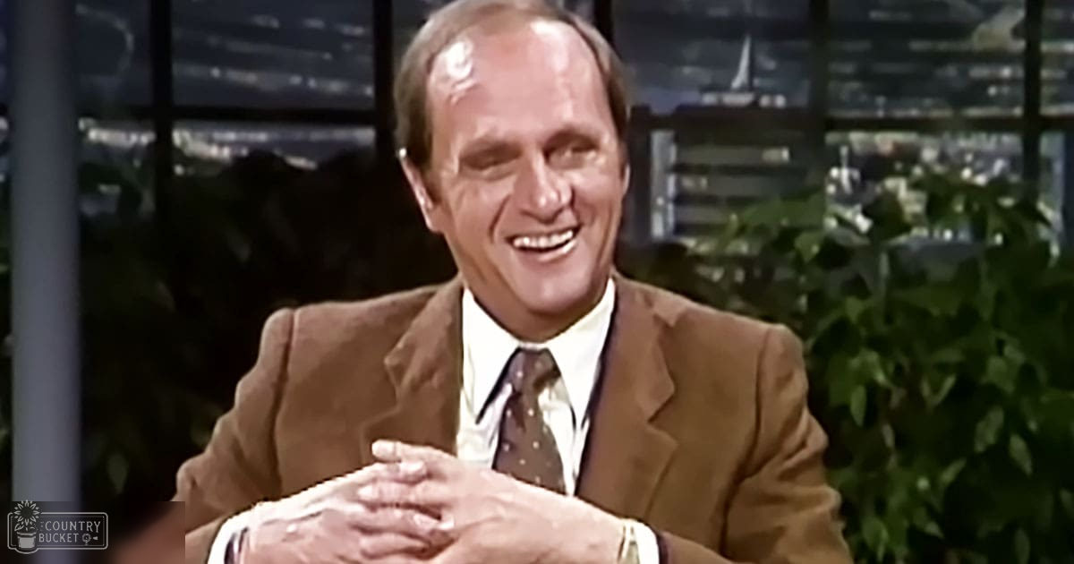 Middle-aged to older adult male wearing a brown suit jacket with white shirt and dark tie with small dots in a studio setting at night.