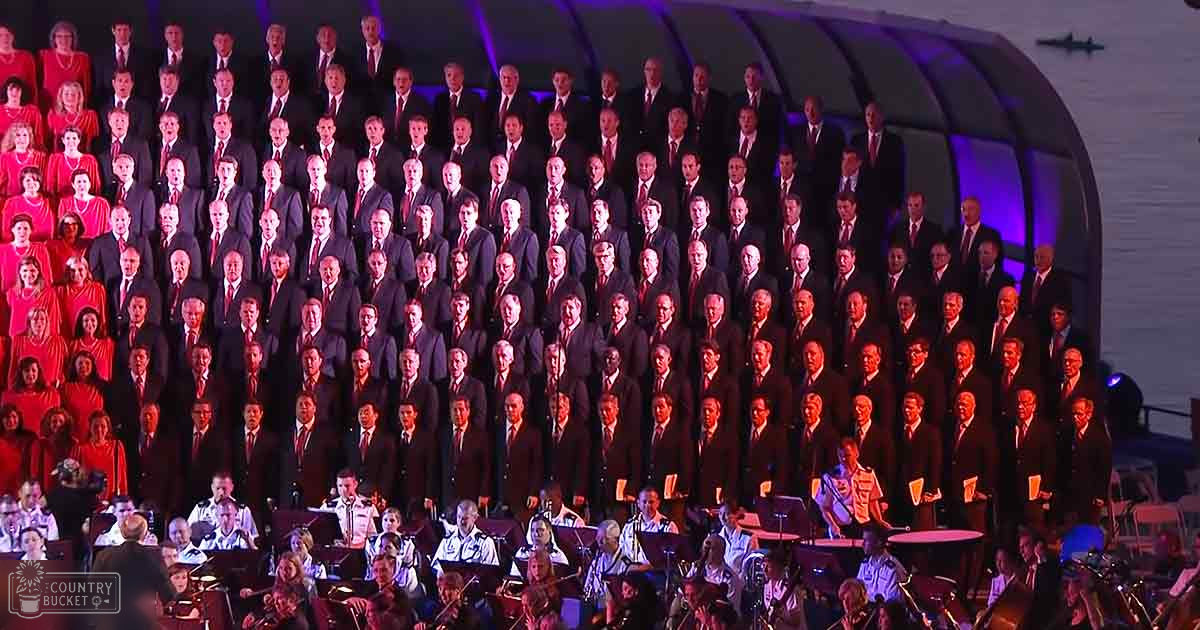 A large choir performing outdoors at night near water, with adults wearing formal attire and red dresses or gowns, set against a backdrop of musical instruments and faint reflections on water in early evening or dusk.