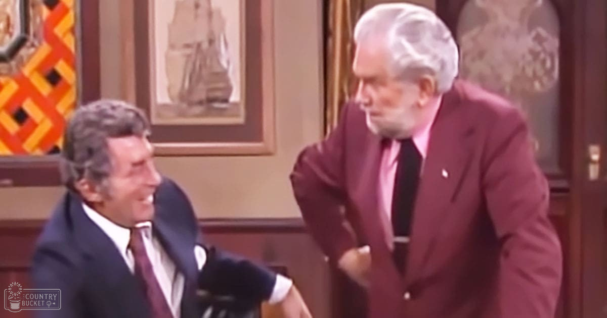 Two middle-aged and older adult males in formal suits interacting humorously indoors with mid-20th century decor, framed ship artwork on the wall.