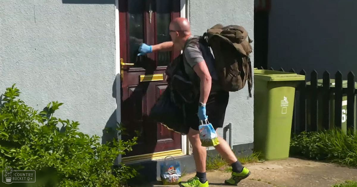 An adult male in a dark vest over a gray t-shirt and black shorts delivers groceries to someone inside a house with white trim around the red door, wearing blue gloves and bright green shoes.