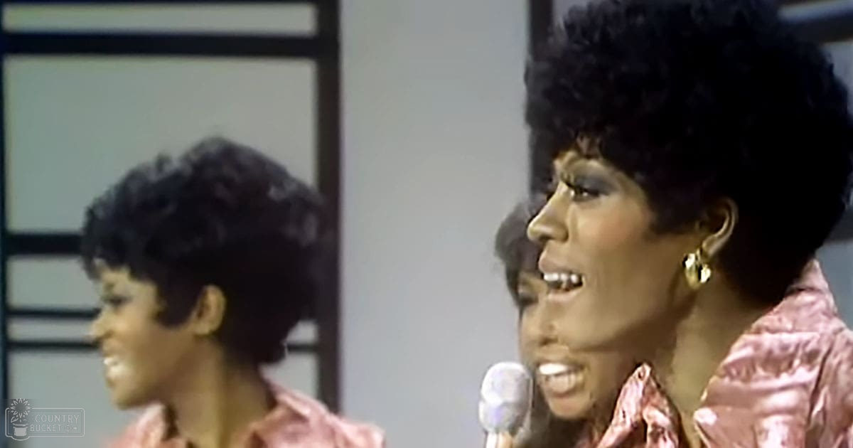 A woman with large hoop earrings, wearing a shiny pink garment in an indoor studio setting. Another individual is partially visible behind her.