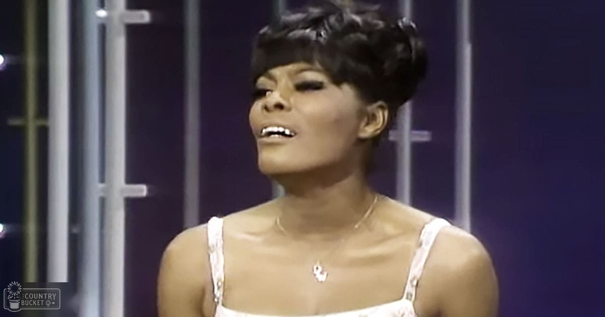 A young adult woman wearing a fitted, white spaghetti strap top with small decorative details on the straps, accessorized with a delicate necklace in an indoor stage or set design environment.