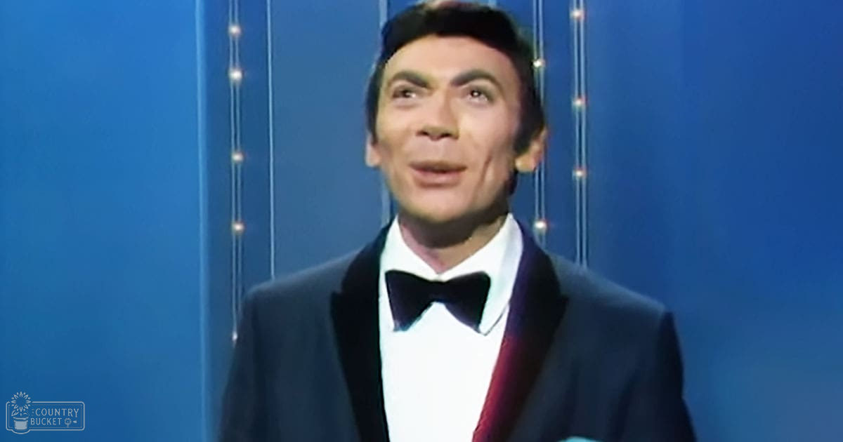 A middle-aged man wearing a classic black tuxedo with a bow tie, standing against a stage-like backdrop with blue vertical lines and small lights.