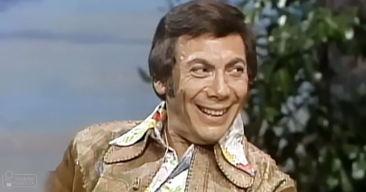 Middle-aged adult male wearing a patterned shirt with a jacket, standing indoors near plants during daytime.