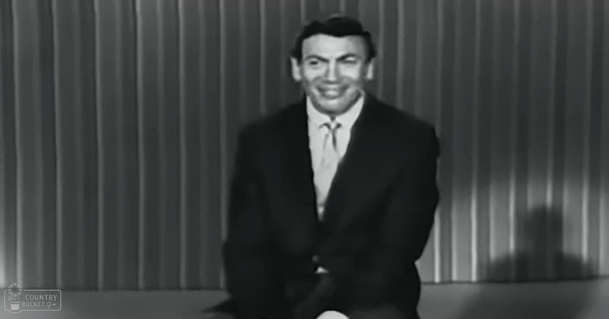 An adult male in a dark suit jacket with a white shirt under an indoor setting, possibly a stage or studio, with vertical blinds and daytime lighting.