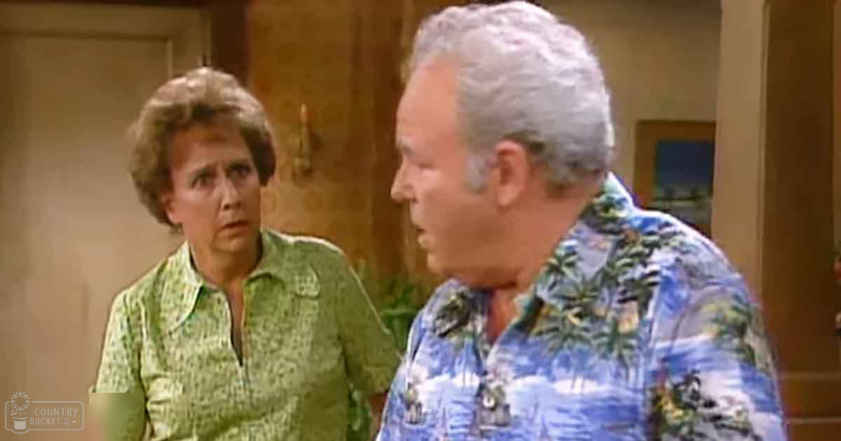 An older adult male wearing a blue Hawaiian-style shirt with palm tree patterns, standing indoors near an older adult female in a green patterned blouse. The setting appears to be a neutral-colored indoor room possibly from an older-style home or apartment.