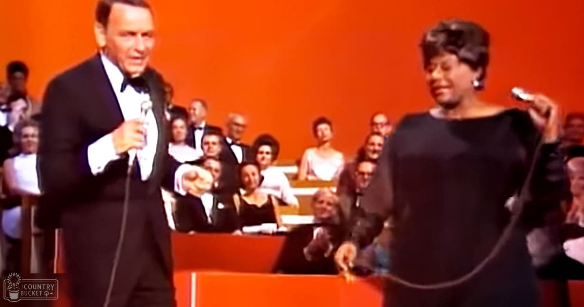 A musical performance scene at an indoor venue with a middle-aged adult male singer in a black tuxedo and a middle-aged adult female performer in an elegant black dress holding a microphone, set against orange walls and wooden benches filled with formally dressed audience members.