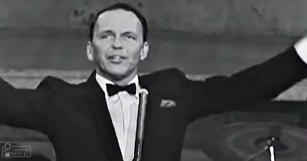 Adult male performer wearing a well-fitted black tuxedo with white shirt and bow tie, standing on an indoor stage with a microphone stand visible to the right.