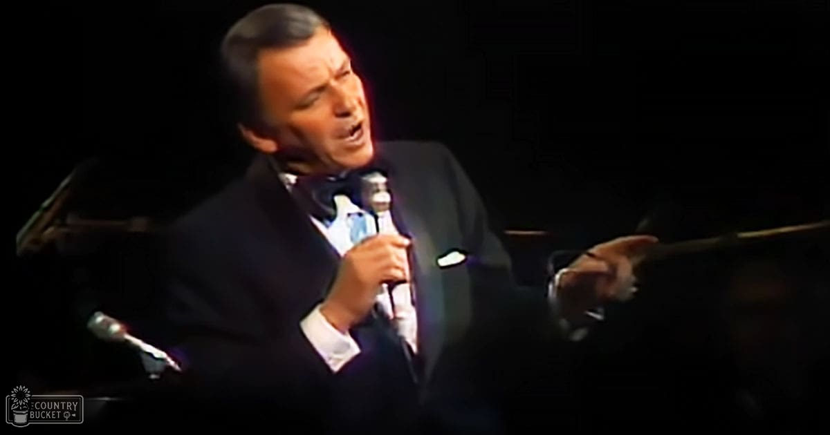 A middle-aged male singer performing on stage, wearing a dark suit jacket with a patterned tie, holding a microphone and sheet music in an indoor concert hall setting.