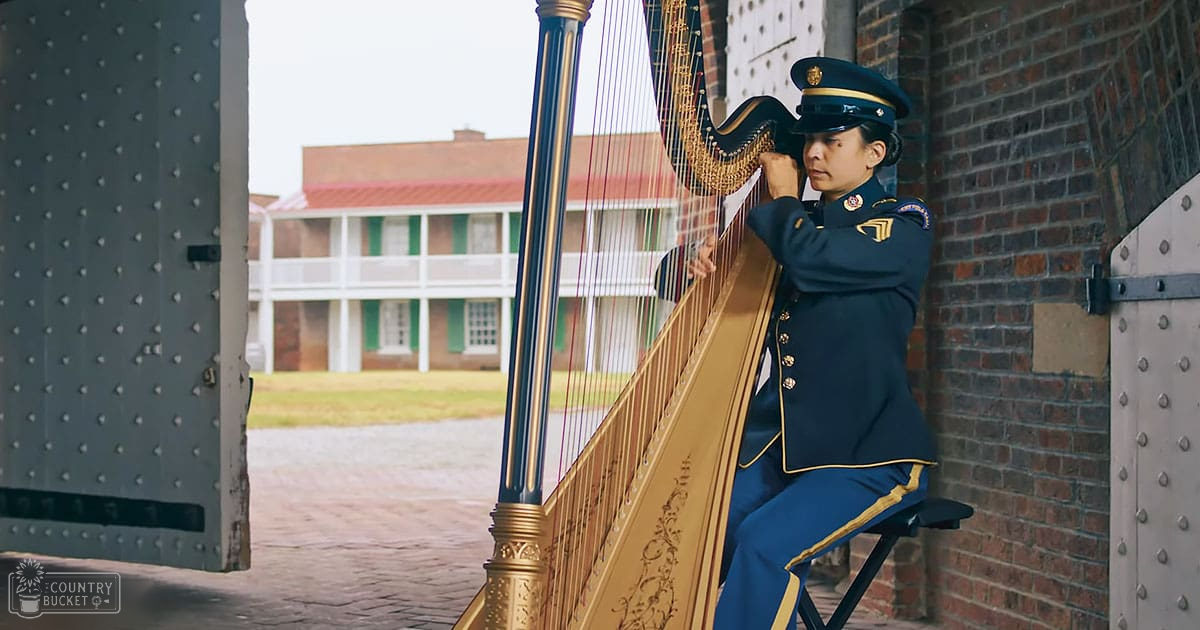 An adult female harpist playing a golden harp in front of Fort McHenry, Baltimore; clear daytime scene with historic fort and white building in background
