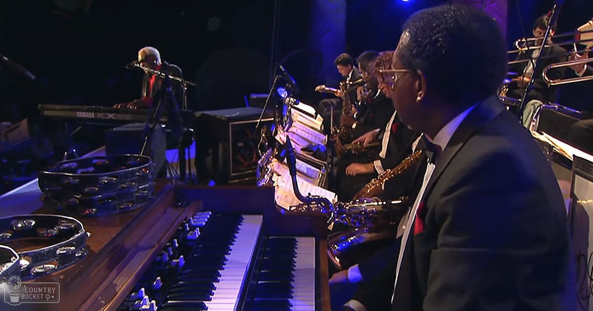 A concert hall performance with an adult male musician playing organ, surrounded by other musicians in formal attire under stage lighting.
