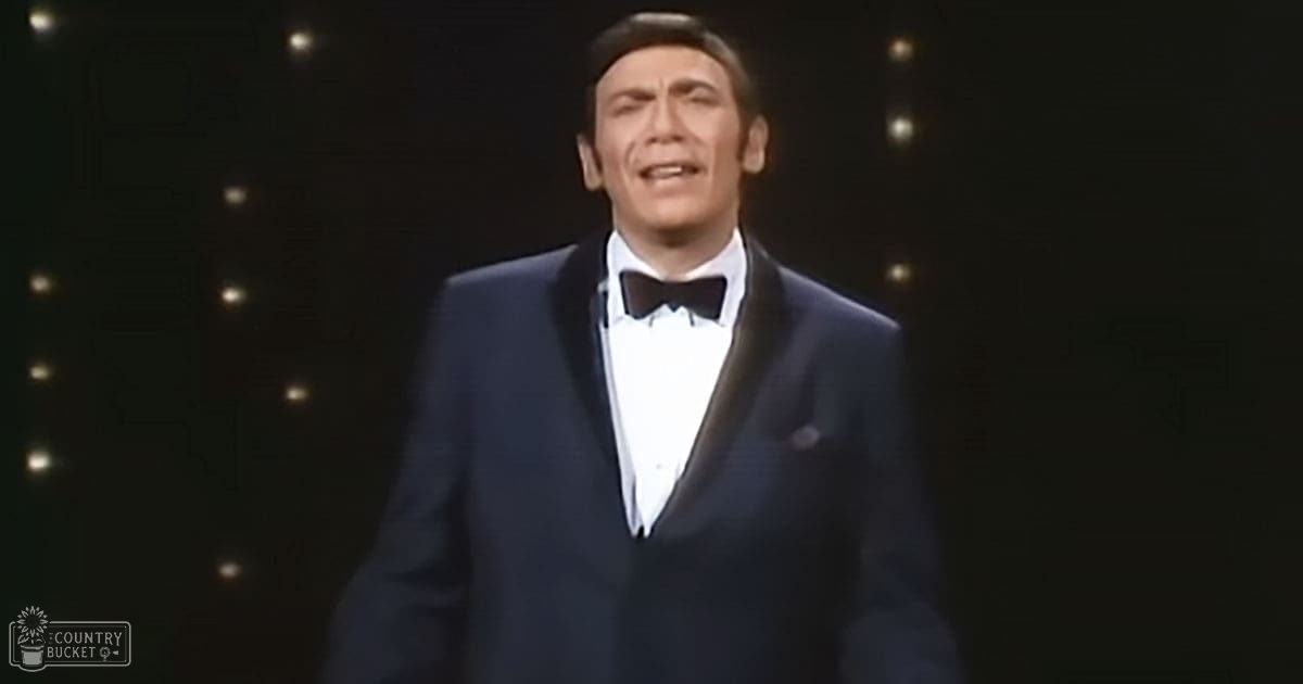 An adult male performer in a dark blue tuxedo with white shirt and black bow tie, standing against a starry background suggesting a stage setting.