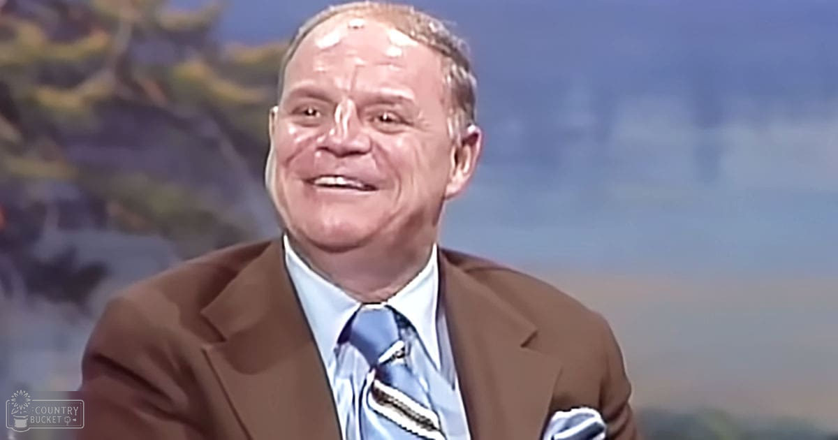 Older adult male in a studio setting with brown blazer and blue dress shirt against an outdoor backdrop depicting mountains and trees under a blue sky during daytime.
