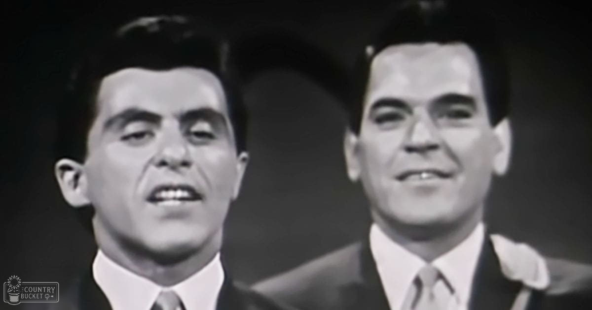 Two adult male singers in suits, one navy or dark, the other light gray or brown, wearing collared shirts and ties, against a studio background with even lighting.