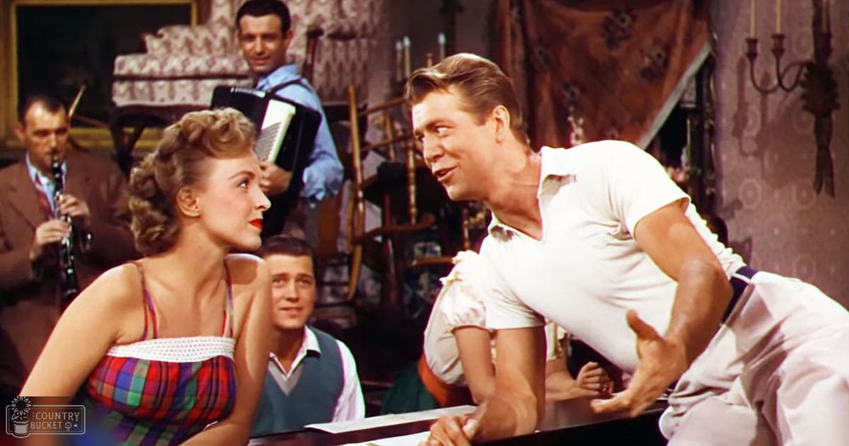 Adult male in a mid-20th-century white shirt interacts with others at an indoor social gathering with patterned decor and daylight.