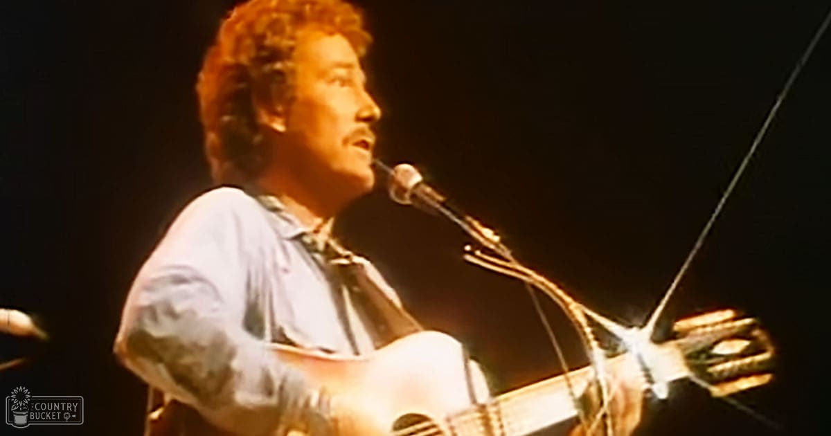 Middle-aged male performer on stage at a concert, wearing a light blue, long-sleeved shirt with rolled-up sleeves, holding an acoustic guitar.