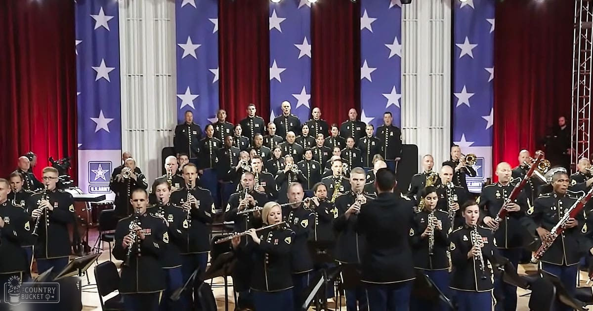 An adult male and female military band performing in front of American flags at an indoor venue with red curtains and bright stage lighting.