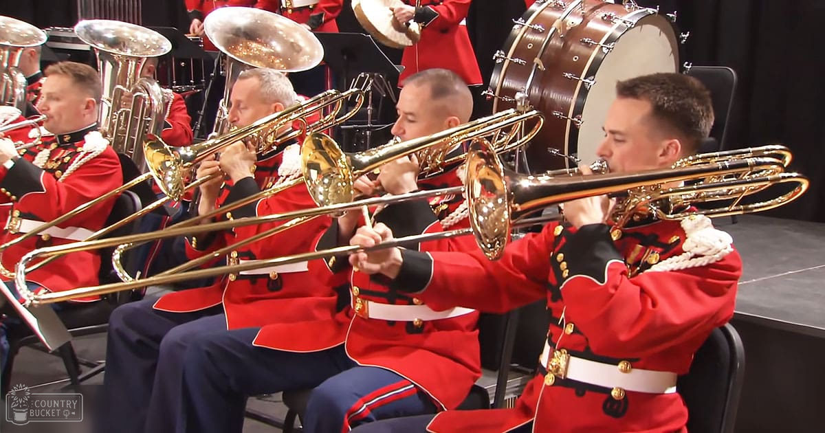 A group of adult male musicians in red military-style uniforms playing trombones indoors, likely at a formal performance venue during winter.