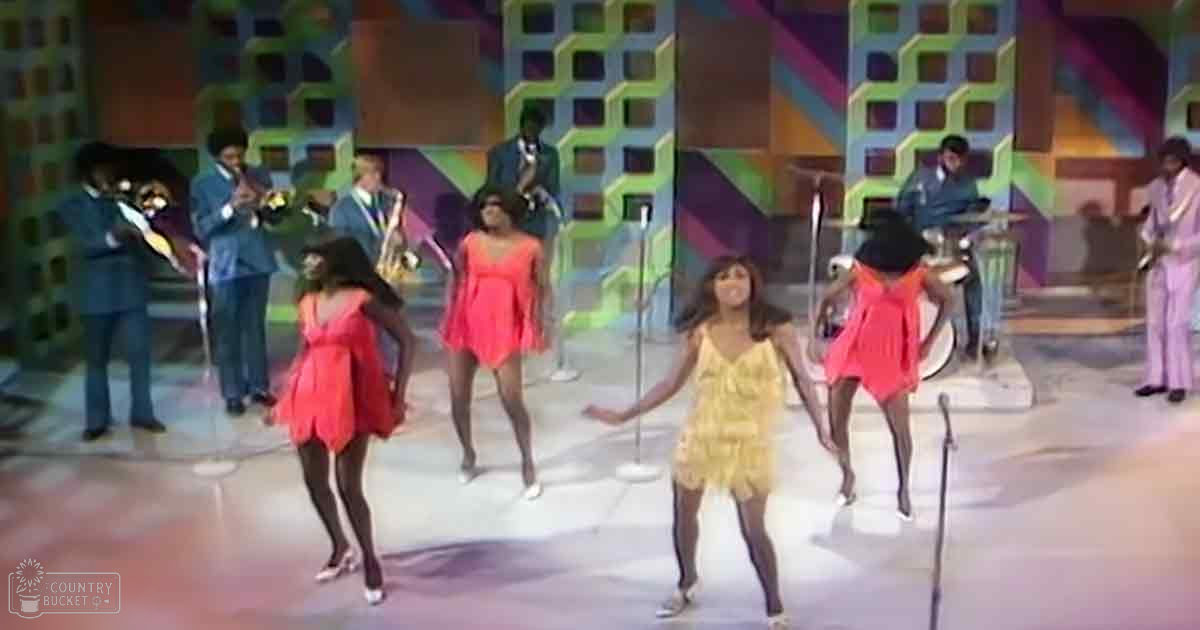 Children in red and yellow dresses perform on stage with adult musicians playing brass instruments, set against a colorful geometric backdrop.