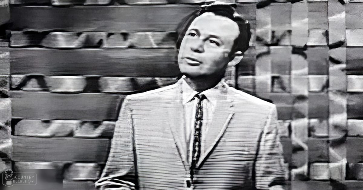Adult male in a light-colored suit jacket and patterned tie, seated on a dark chair or bench against a brick wall backdrop, likely in an indoor studio or theater setting.