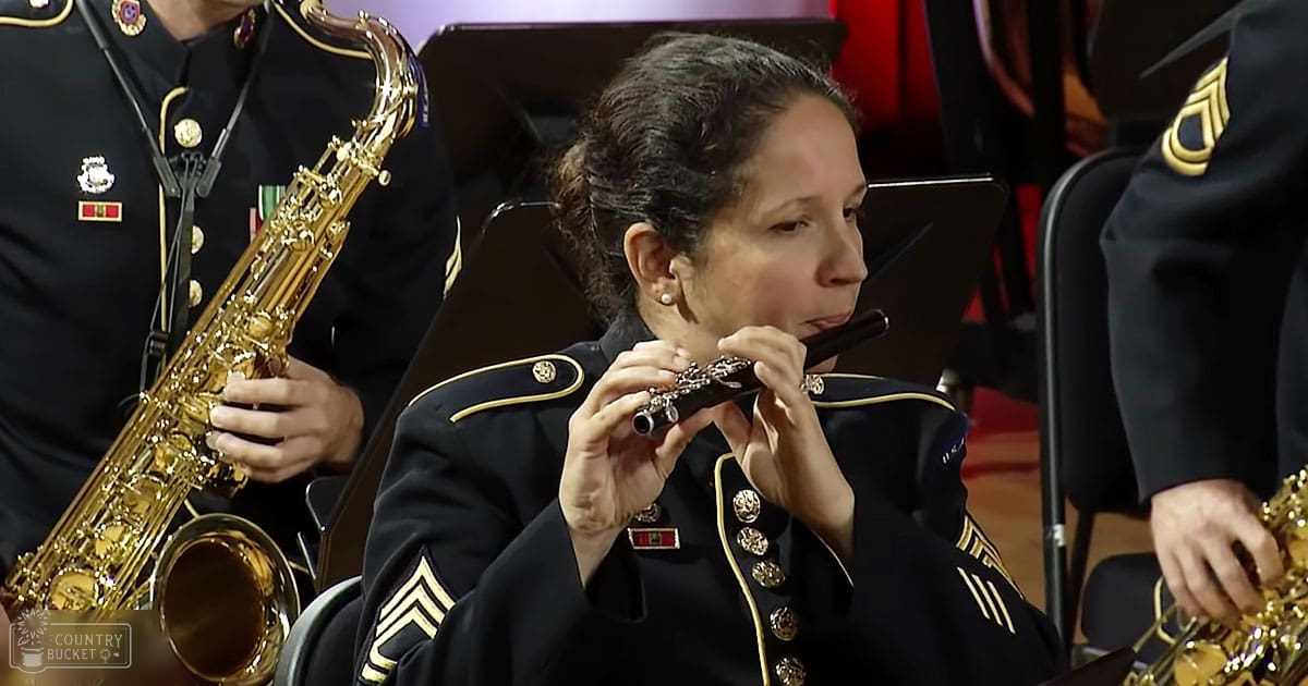 An adult female musician plays a flute indoors with another person holding a saxophone nearby; setting appears to be an evening concert hall or auditorium, with dark blue jackets and gold accents visible.