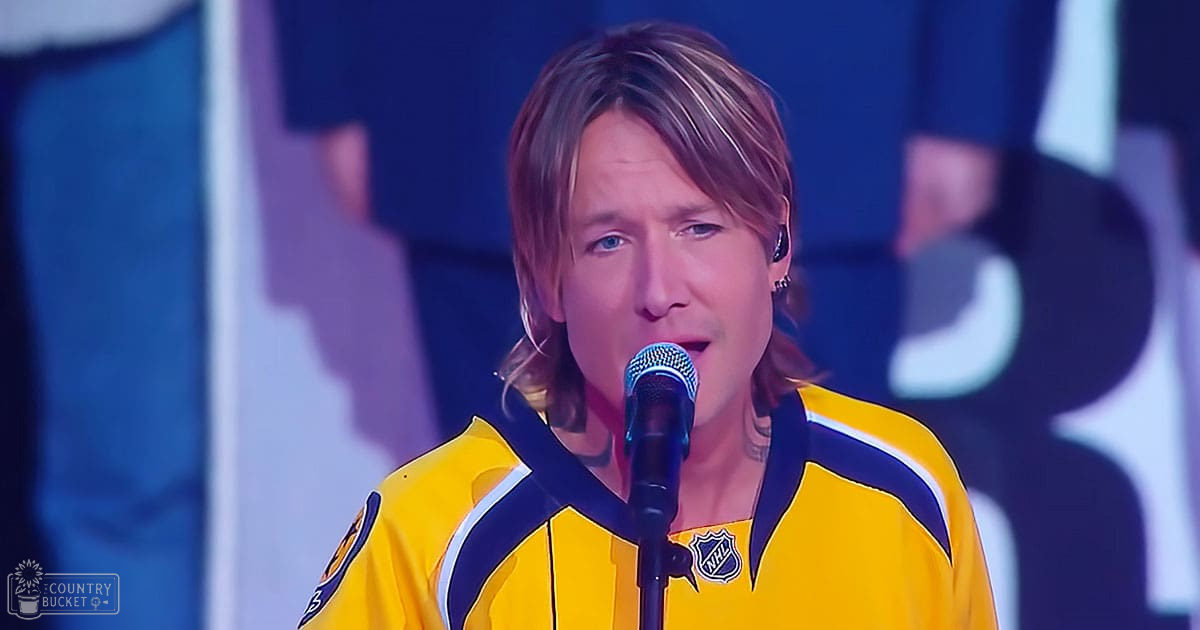 A middle-aged male performer in a yellow and blue hockey jersey holding a microphone, likely at an indoor sports-related event with stage lighting.