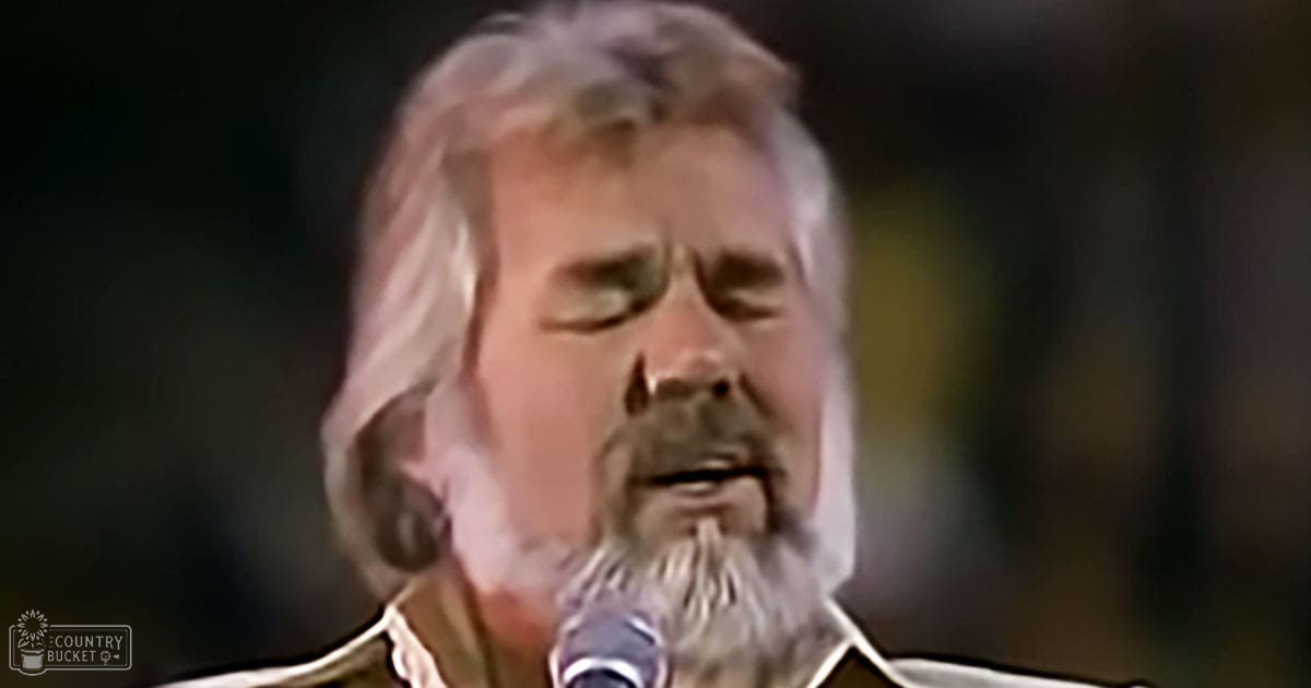 A middle-aged male singer wearing a dark, textured garment with a light-colored collar performs into a microphone at an indoor event under dim stage lighting.