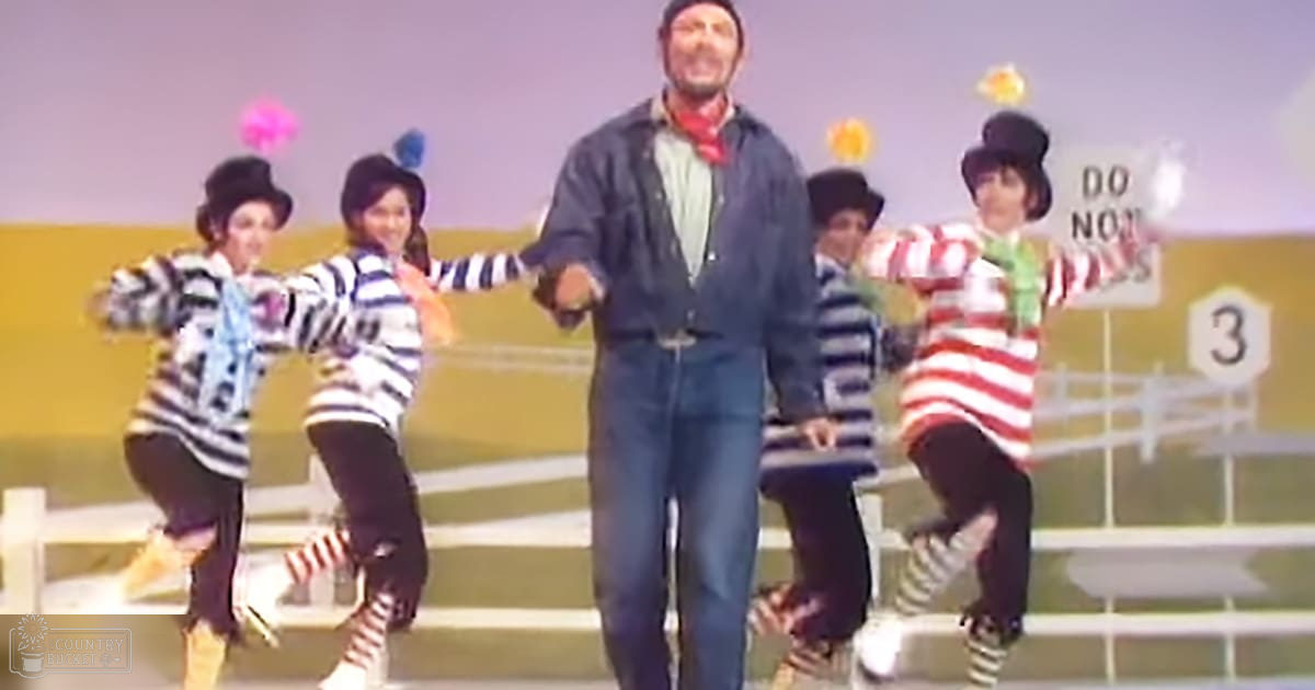 A middle-aged man in a denim jacket outdoors with young adults wearing striped tops and pants, near a white picket fence under bright daylight.
