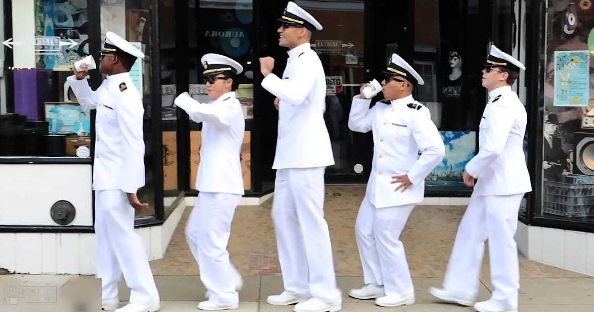 Group of five young adult male sailors in white uniforms, possibly part of a performance or event, outdoors with urban storefronts in the background.