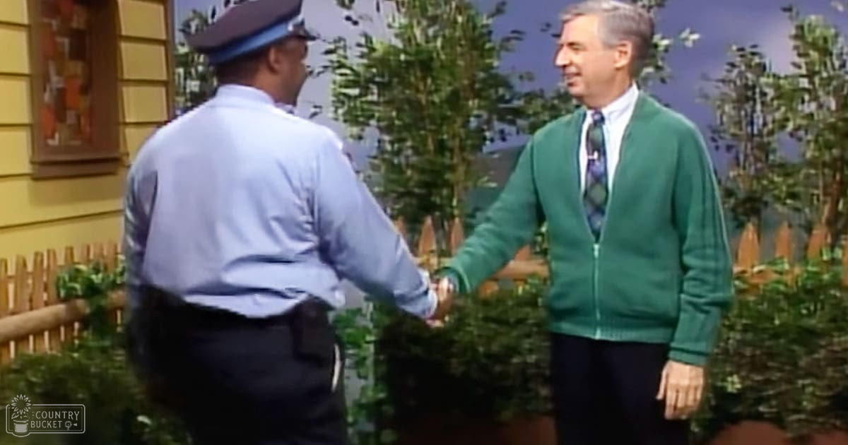 Two middle-aged men interacting outdoors; one wearing a green cardigan and tie, the other in a blue uniform shirt with a hat.