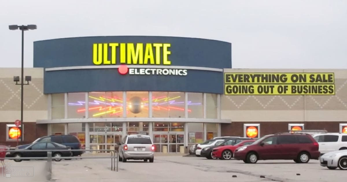 An electronics store with various vehicles parked outside, signage indicating 'Ultimate Electronics' on sale.