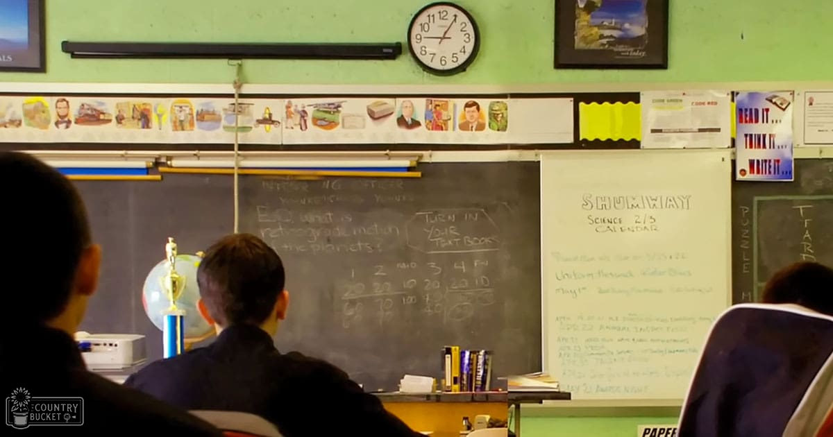 A classroom scene with children and adult female teacher, educational posters, globe, calendar titled "Shumway Science 2/3 Calendar", natural or fluorescent light, green walls, blackboard, white clock face, blue trophy indicating academic achievement.
