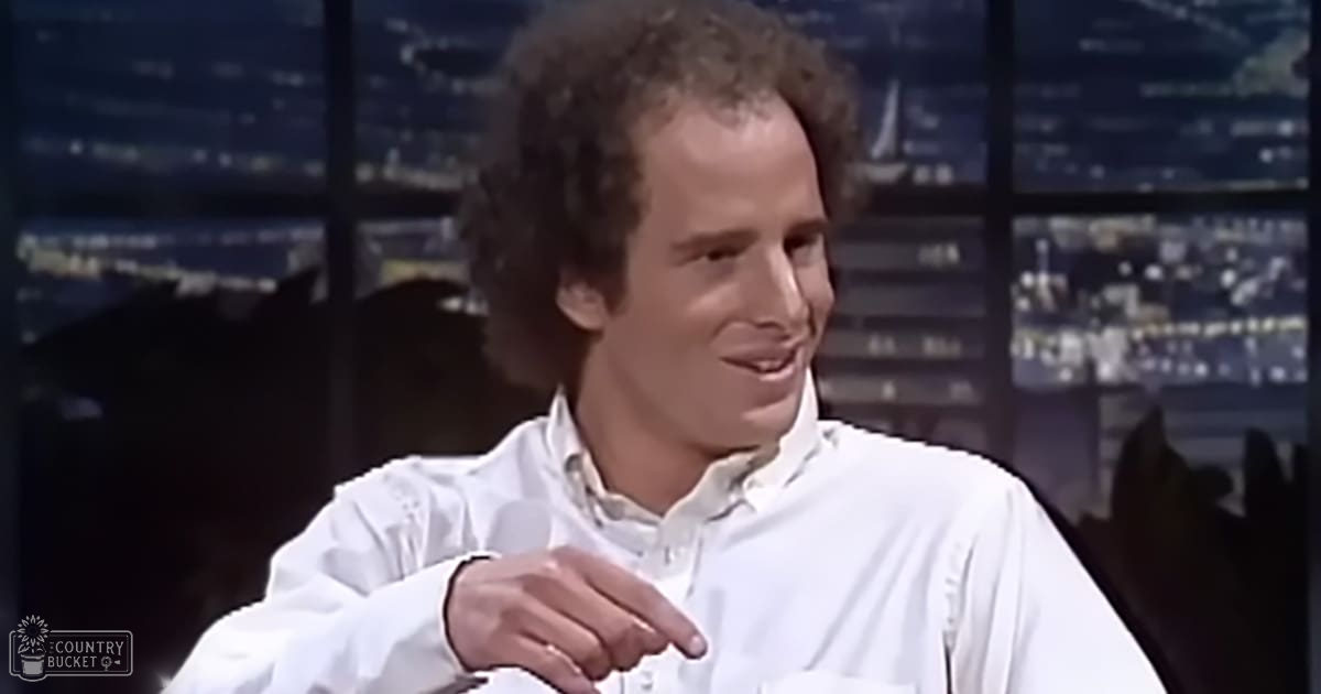 Young adult male wearing a relaxed fit, white button-up shirt with long sleeves slightly rolled up, set against an indoor studio or talk show backdrop at night with illuminated cityscape in the background.
