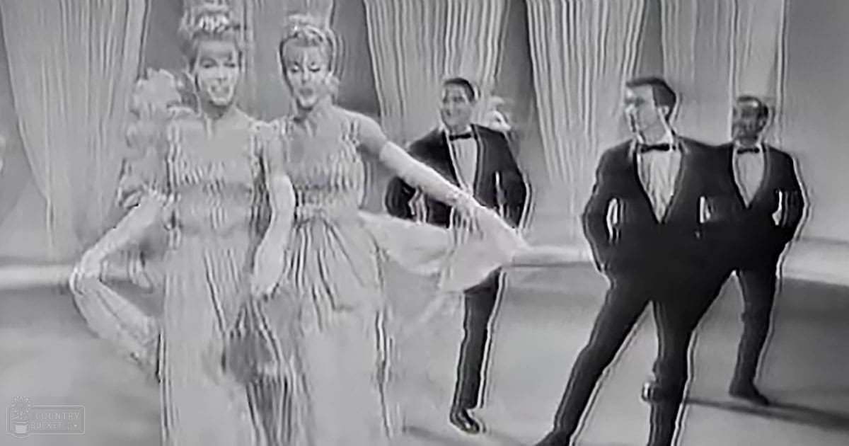A dance scene with individuals dressed in frilly dresses and tuxedos set against heavy striped curtains, suggesting an indoor theater or studio environment.