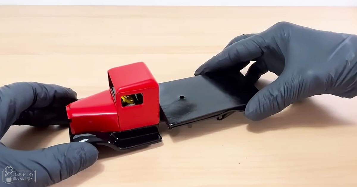 A close-up of a red and black toy truck being held by dark gray, latex or nitrile-gloved hands on an indoor light wooden surface.