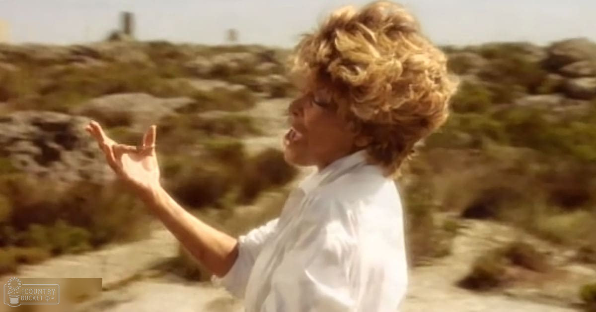 A young adult male with curly blonde hair stands outdoors in a desert-like environment under clear midday summer or spring sky.