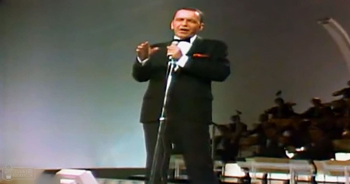 A middle-aged male performer singing on stage in a concert hall, wearing a dark suit with white shirt and black bow tie, surrounded by an orchestra under dramatic lighting.