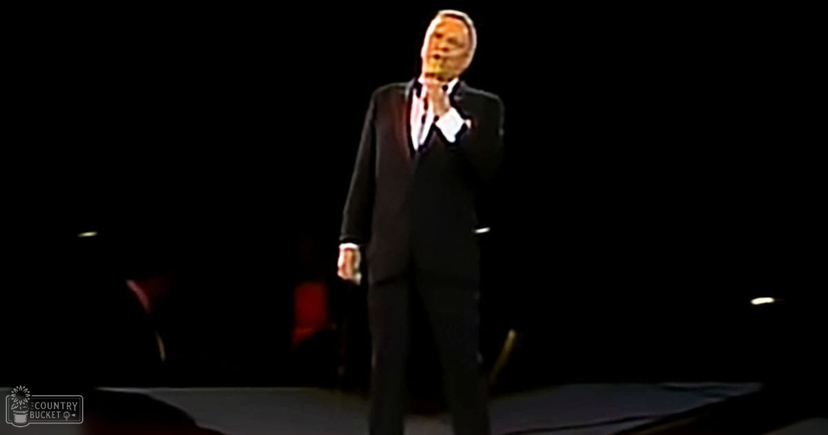 A male adult singer dressed in a dark mid-20th-century style suit, performing on stage under dim lighting with faint outlines of chairs and a dark background.