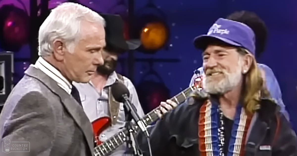 An older adult male singer wearing a gray blazer over a white collared shirt with a dark tie, holding an electric guitar on stage with another older adult male in a blue baseball cap and black jacket with red accents, both also holding guitars. The setting is indoors at a concert venue.