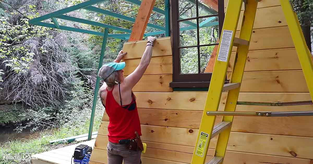 A woman in a red sleeveless top and gray pants works on a wooden structure near a river, surrounded by greenery under natural daylight.